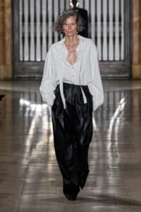Model walking a fashion runway in a white pin-striped blouse paired with black wide-leg pants, hands in pockets.