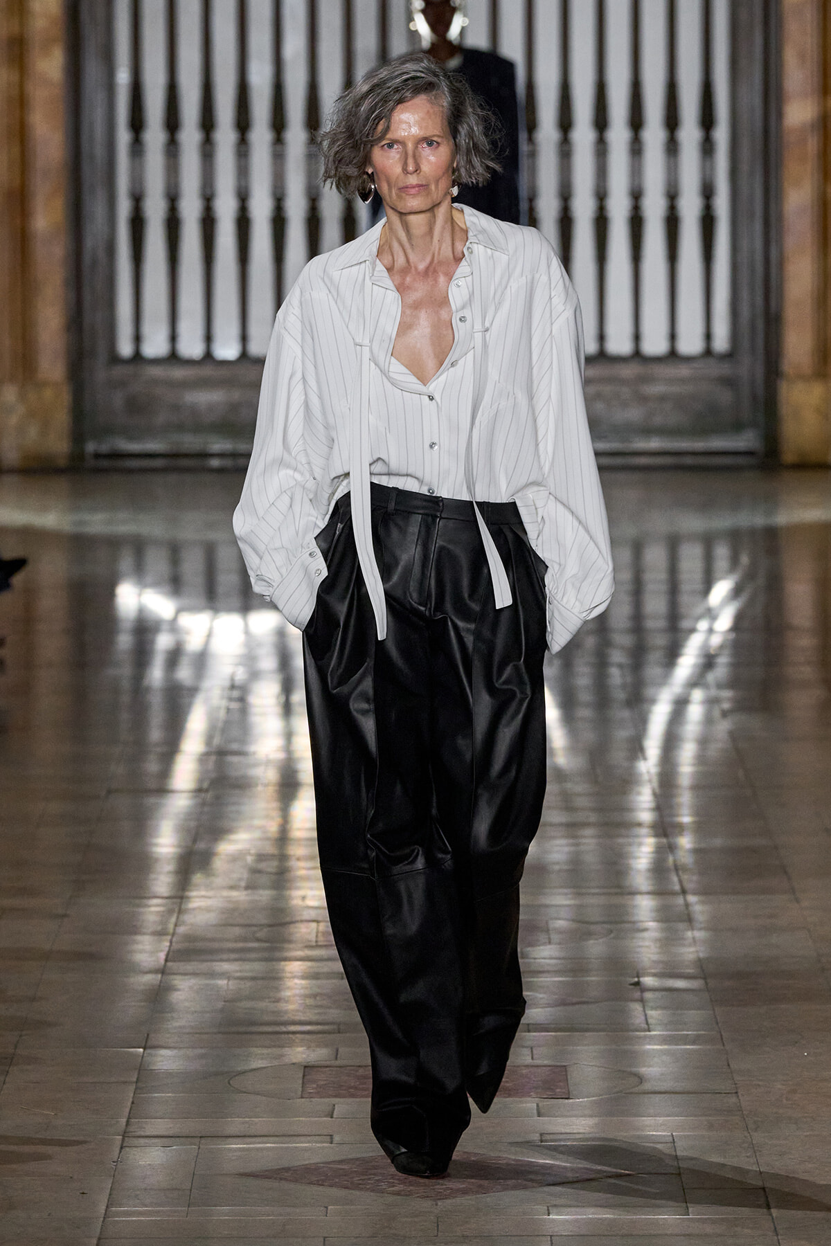 Model walking a fashion runway in a white pin-striped blouse paired with black wide-leg pants, hands in pockets.