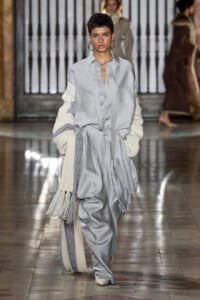 Model walking a runway in a loose gray shirt-dress and matching wide trousers, layered with a cream-knit scarf draped over her arm.