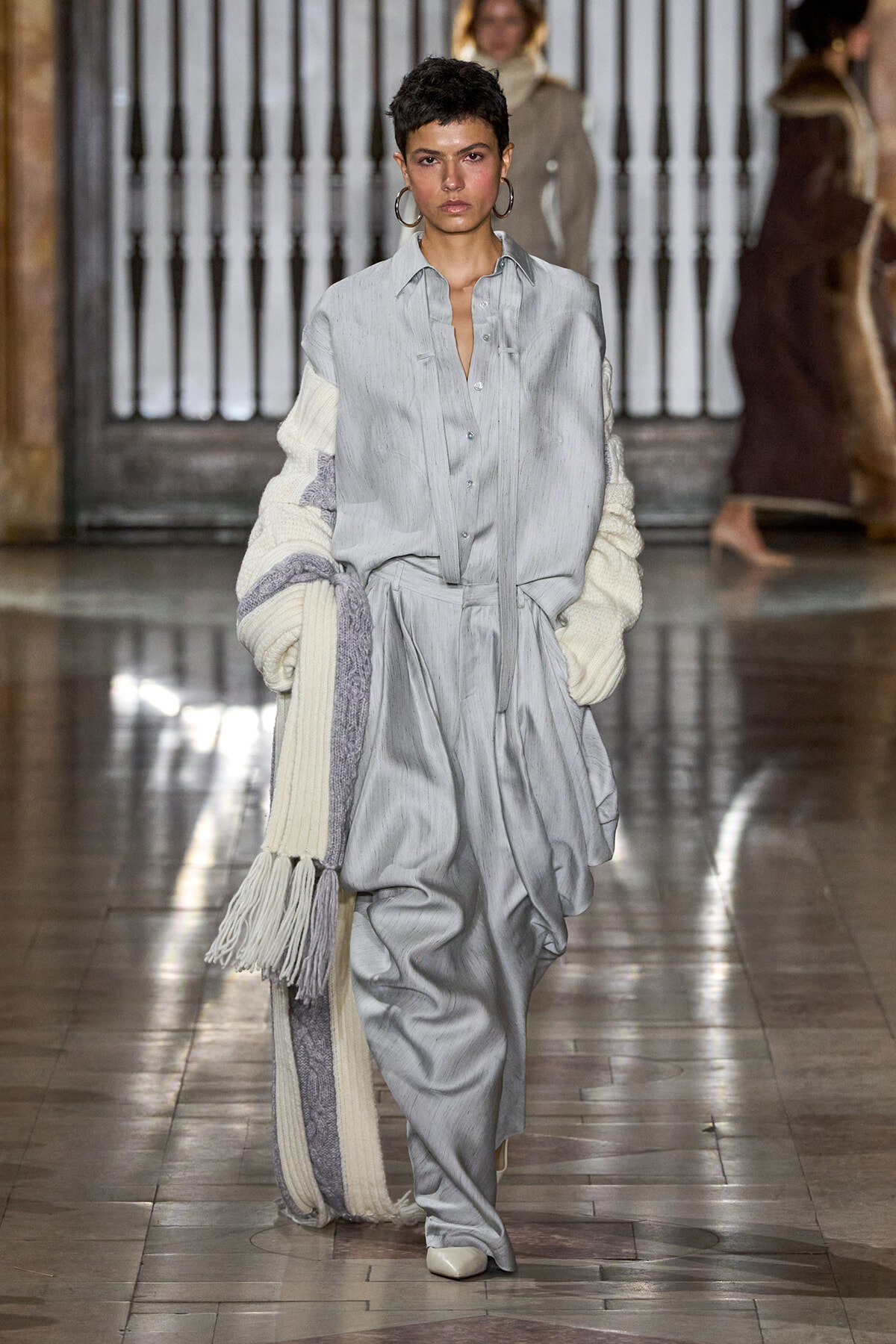 Model walking a runway in a loose gray shirt-dress and matching wide trousers, layered with a cream-knit scarf draped over her arm.