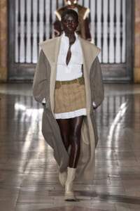 Model walks a runway in a long beige coat over a white blouse and tan plaid mini skirt with beige boots.