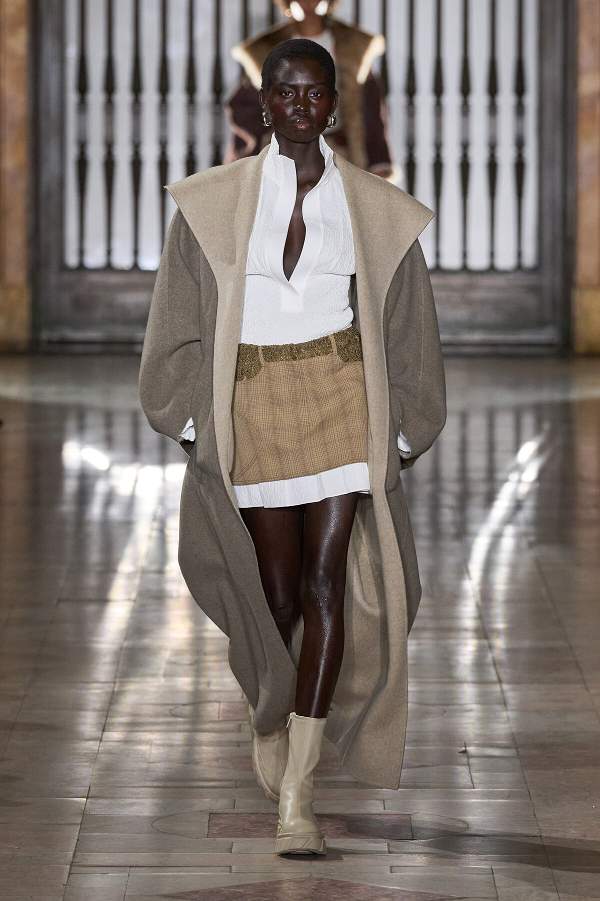 Model walks a runway in a long beige coat over a white blouse and tan plaid mini skirt with beige boots.
