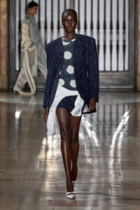 Model walking runway in a navy pinstripe blazer over a gray polka-dot top with a white, wrapped asymmetrical skirt and white heels.