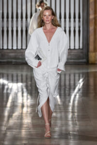 Female model walks a runway in a white pinstripe shirt dress, holding a small booklet and wearing hoop earrings.