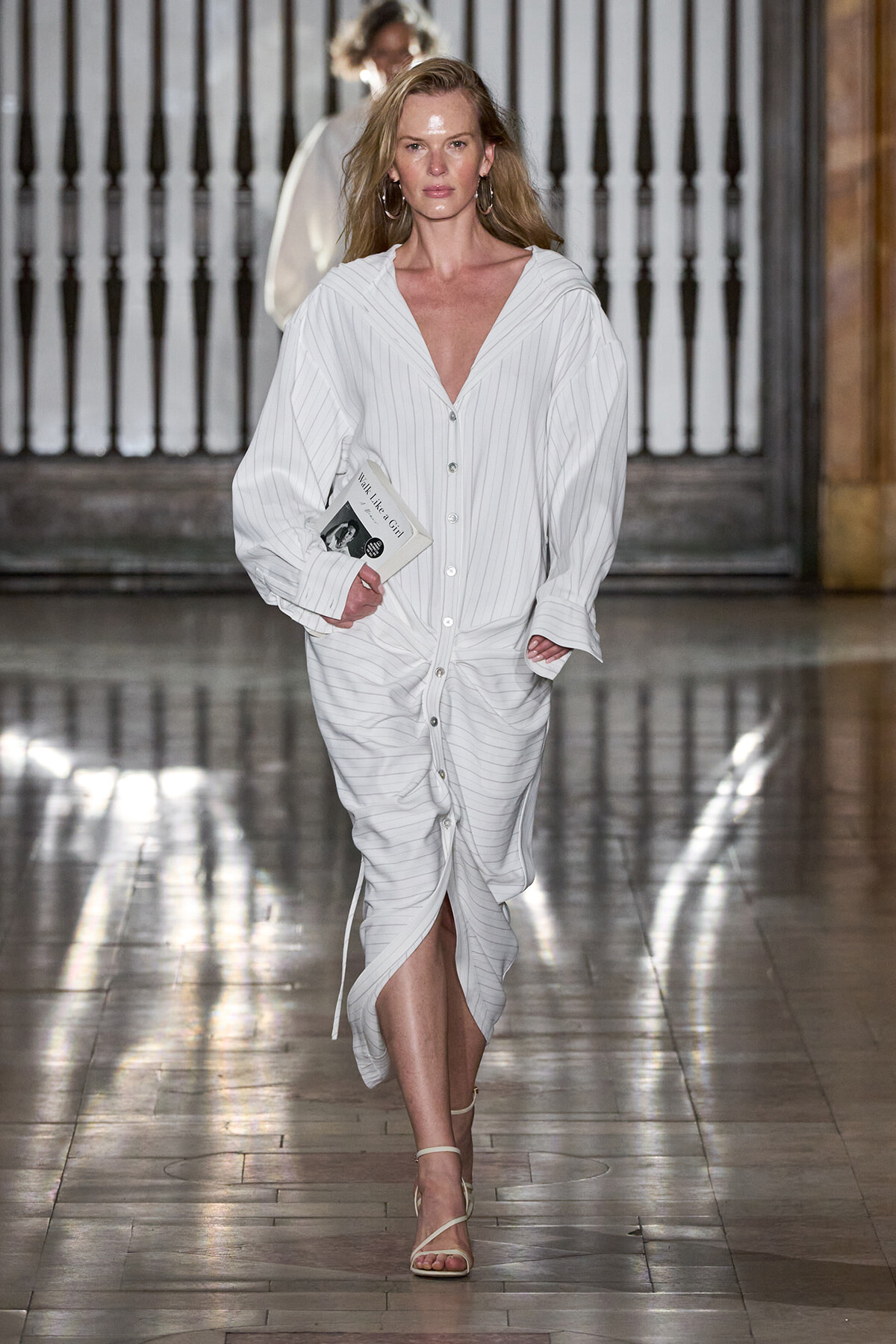Female model walks a runway in a white pinstripe shirt dress, holding a small booklet and wearing hoop earrings.