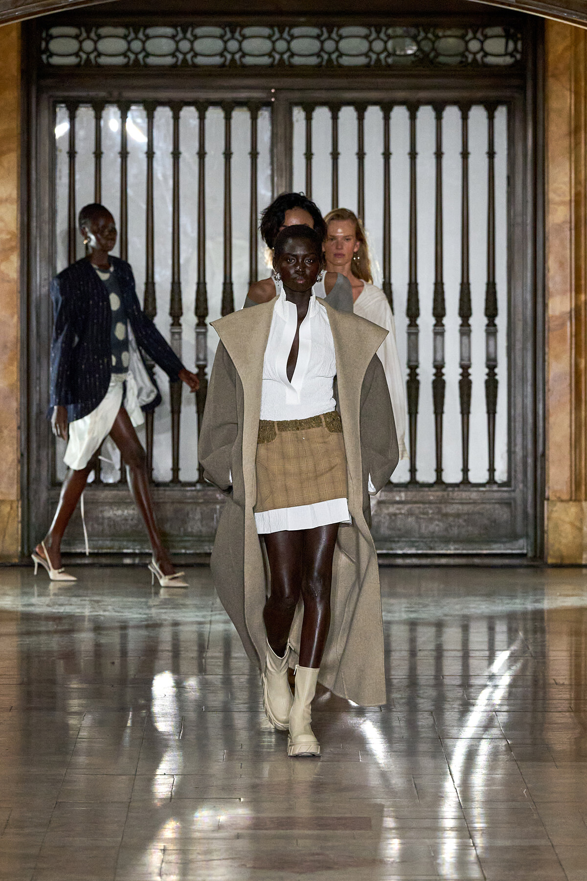 Model walks down a marble hallway wearing a long beige cape over a white blouse and a tan plaid mini skirt, paired with light boots; a fashion show vibe.