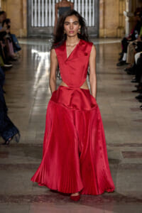 Female runway model wearing a sleeveless red dress with a structured bodice and a wide pleated skirt on a fashion show runway.