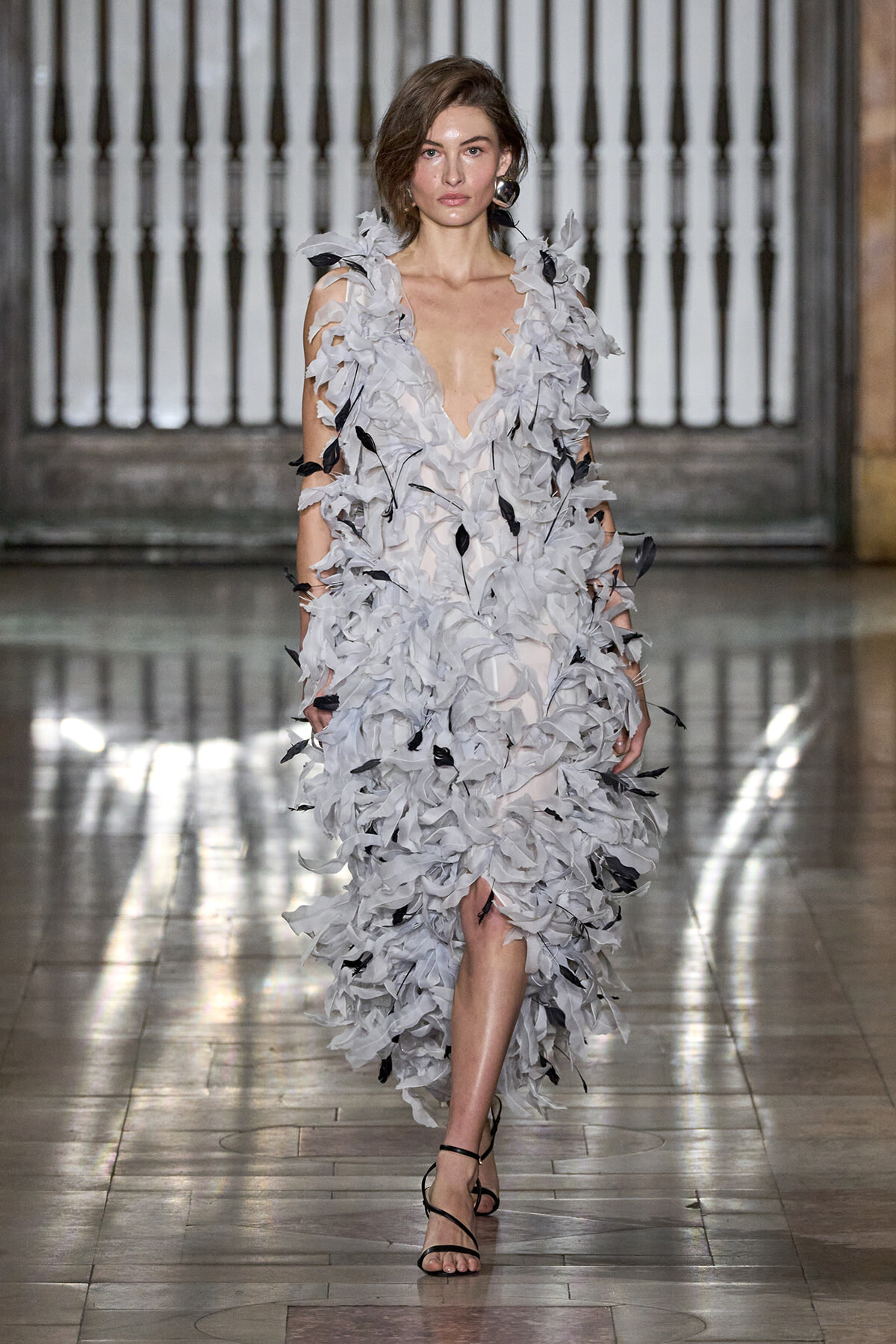 Model walks the runway in a pale gray, leaf-embellished gown with a plunging neckline and black strappy heels in a grand venue.