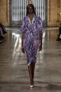 Model walks the runway in a purple floral knee-length dress with mint-green lapels and pink fringe detailing at the hem.