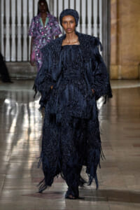 Model walking the runway in a voluminous navy textured fringe gown with a matching head wrap.