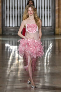 Model walking a runway in a pink, ruffled dress with a sheer bodice and metallic flats; fashion show setting in the background.