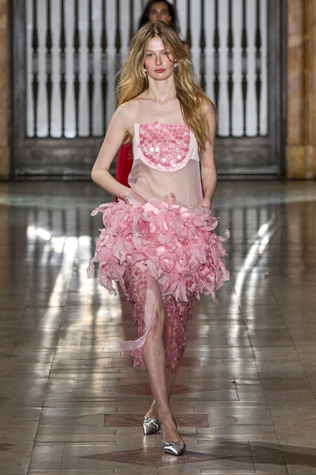 Model walking a runway in a pink, ruffled dress with a sheer bodice and metallic flats; fashion show setting in the background.