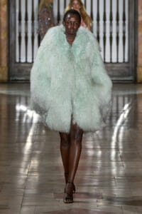 Model walks the runway wearing a voluminous mint-green faux fur coat and black strappy sandals in a fashion show setting.