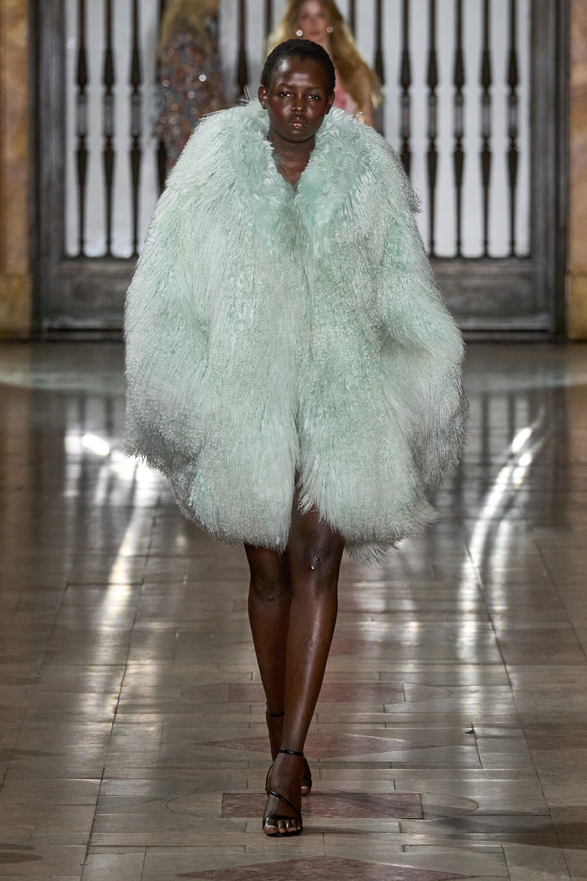 Model walks the runway wearing a voluminous mint-green faux fur coat and black strappy sandals in a fashion show setting.