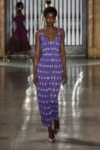 Model walks the runway in a sleeveless purple jumpsuit with white polka dots and a cinched waist, paired with metallic heels.
