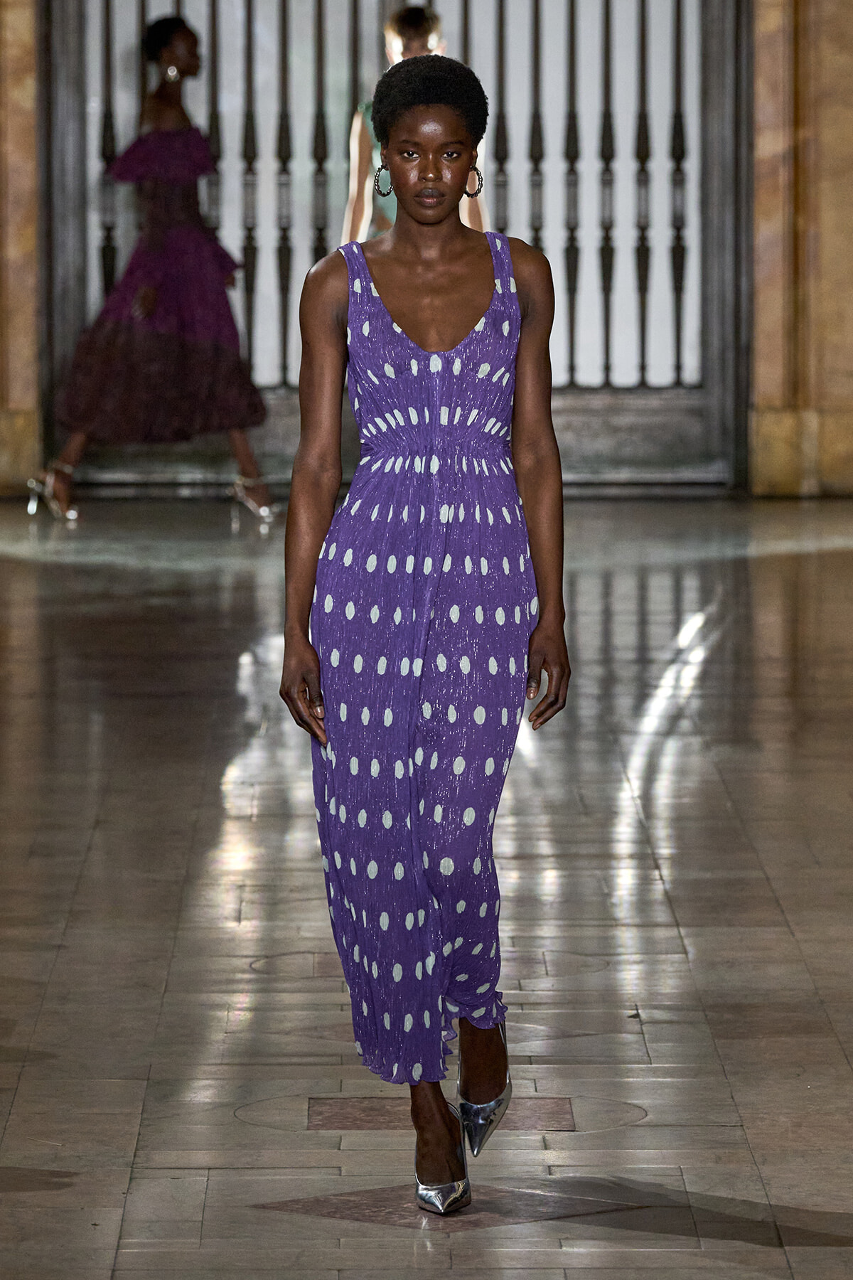 Model walks the runway in a sleeveless purple jumpsuit with white polka dots and a cinched waist, paired with metallic heels.