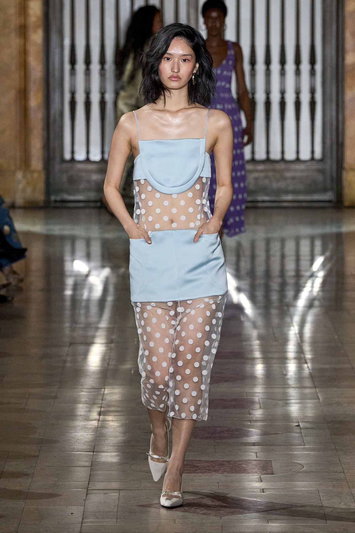 Model walking the runway in a pale blue sleeveless top with a sheer white polka-dot overlay and matching skirt/overlay.