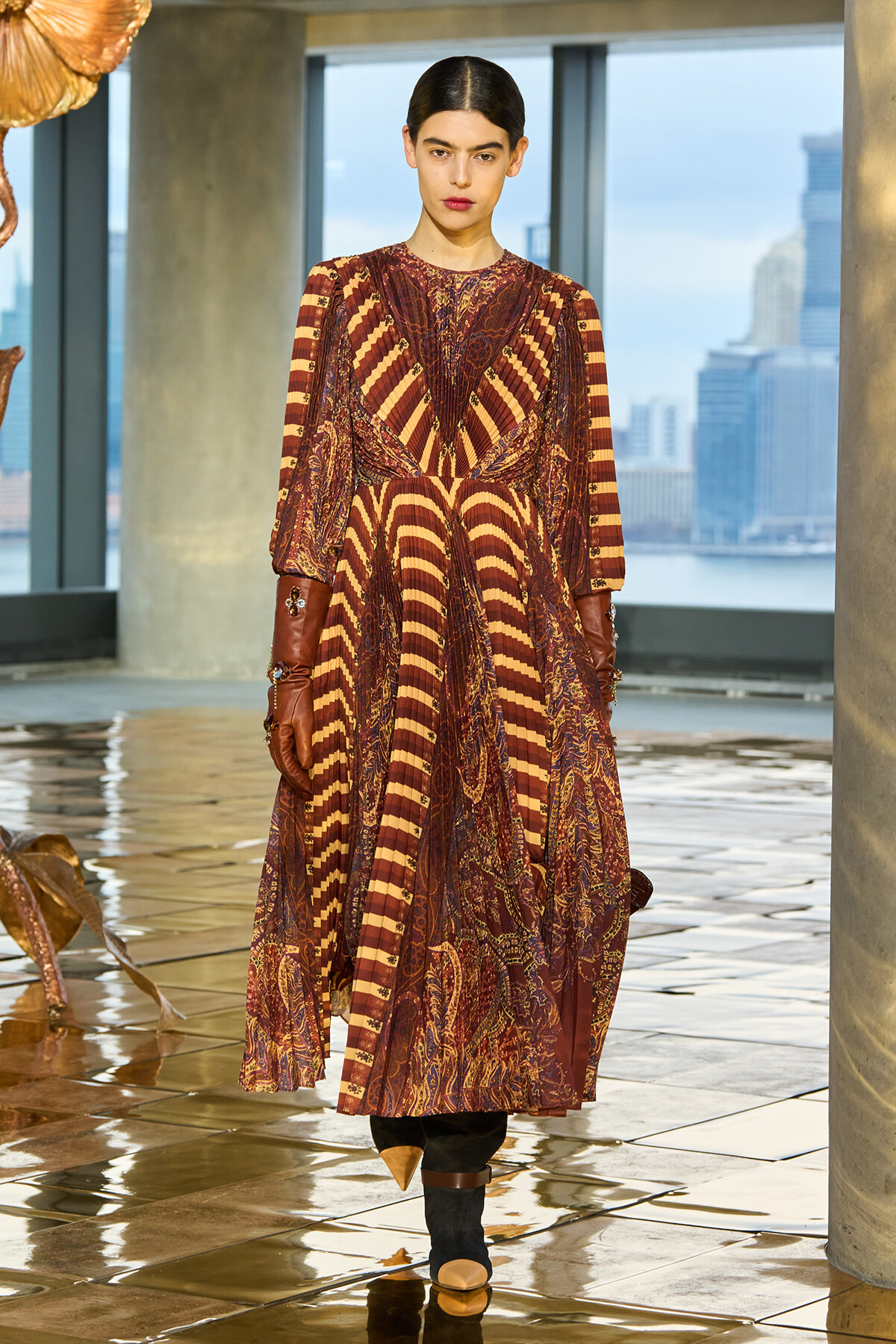 Model wearing a maroon patterned maxi dress with striped panels and brown gloves indoors, city skyline visible through windows behind her.