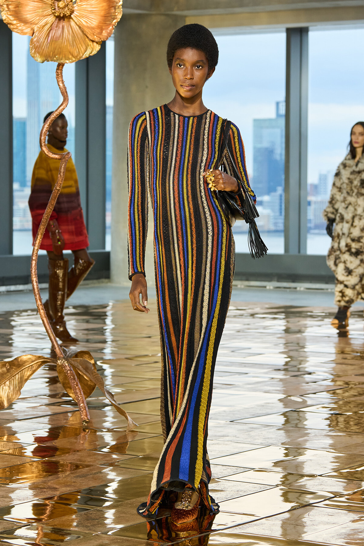 Runway model wears a long multicolored striped knit gown, carrying a black fringe handbag during a fashion show.