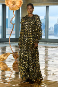 Model wearing a long black dress with gold polka dots and ruffled sleeves, standing on a reflective gold floor beside a large metallic flower sculpture in a skyline setting.