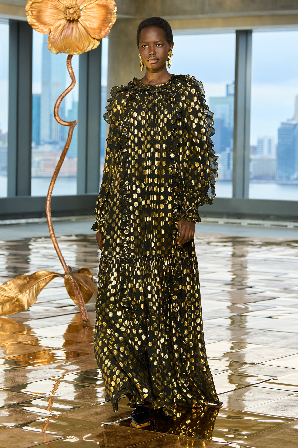 Model wearing a long black dress with gold polka dots and ruffled sleeves, standing on a reflective gold floor beside a large metallic flower sculpture in a skyline setting.