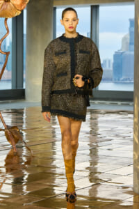 Model wearing a gold-and-black tweed jacket and matching mini skirt with black trim, mustard socks, and strappy sandals, holding a tassel bag.