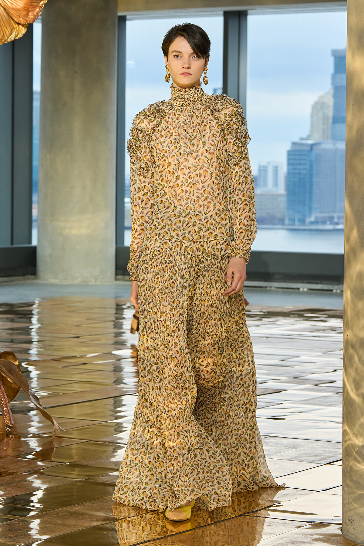 Model wearing a beige floral-print, high-neck smocked dress with long sleeves, standing in a modern indoor space with city skyline beyond the windows.