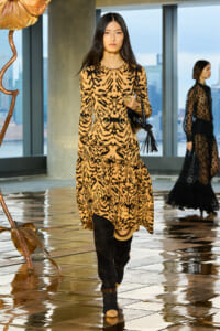 Model wearing a leopard-print dress and knee-high black boots walks the indoor runway, carrying a black fringe bag.