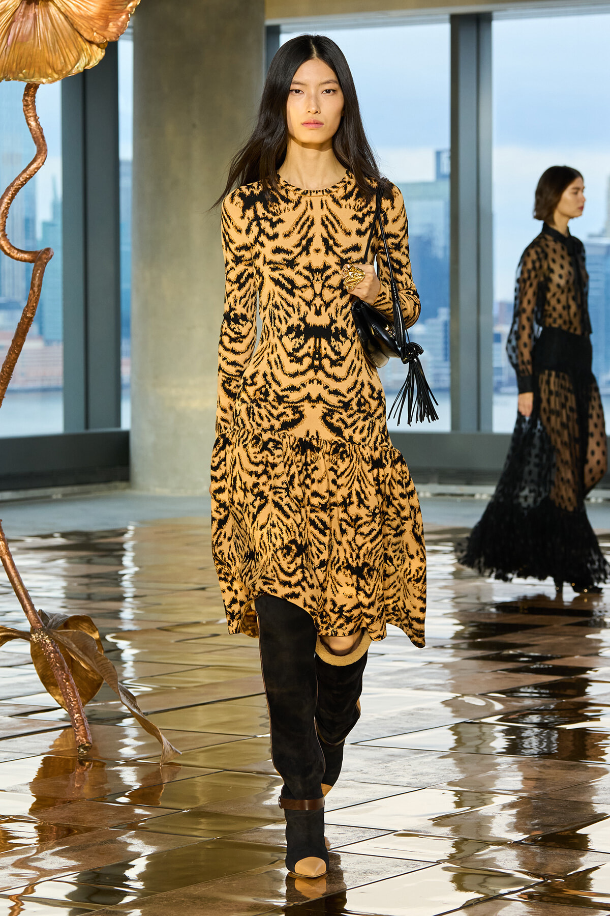 Model wearing a leopard-print dress and knee-high black boots walks the indoor runway, carrying a black fringe bag.