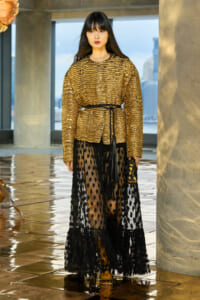 Model wearing a metallic gold textured blouse belted at the waist, paired with a sheer black polka-dot skirt and a small clutch.