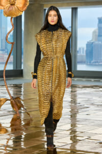 Model wearing a gold textured sleeveless coat over a black turtleneck and pants, bronze boots, walking on a reflective tiled floor with a city skyline in the background.