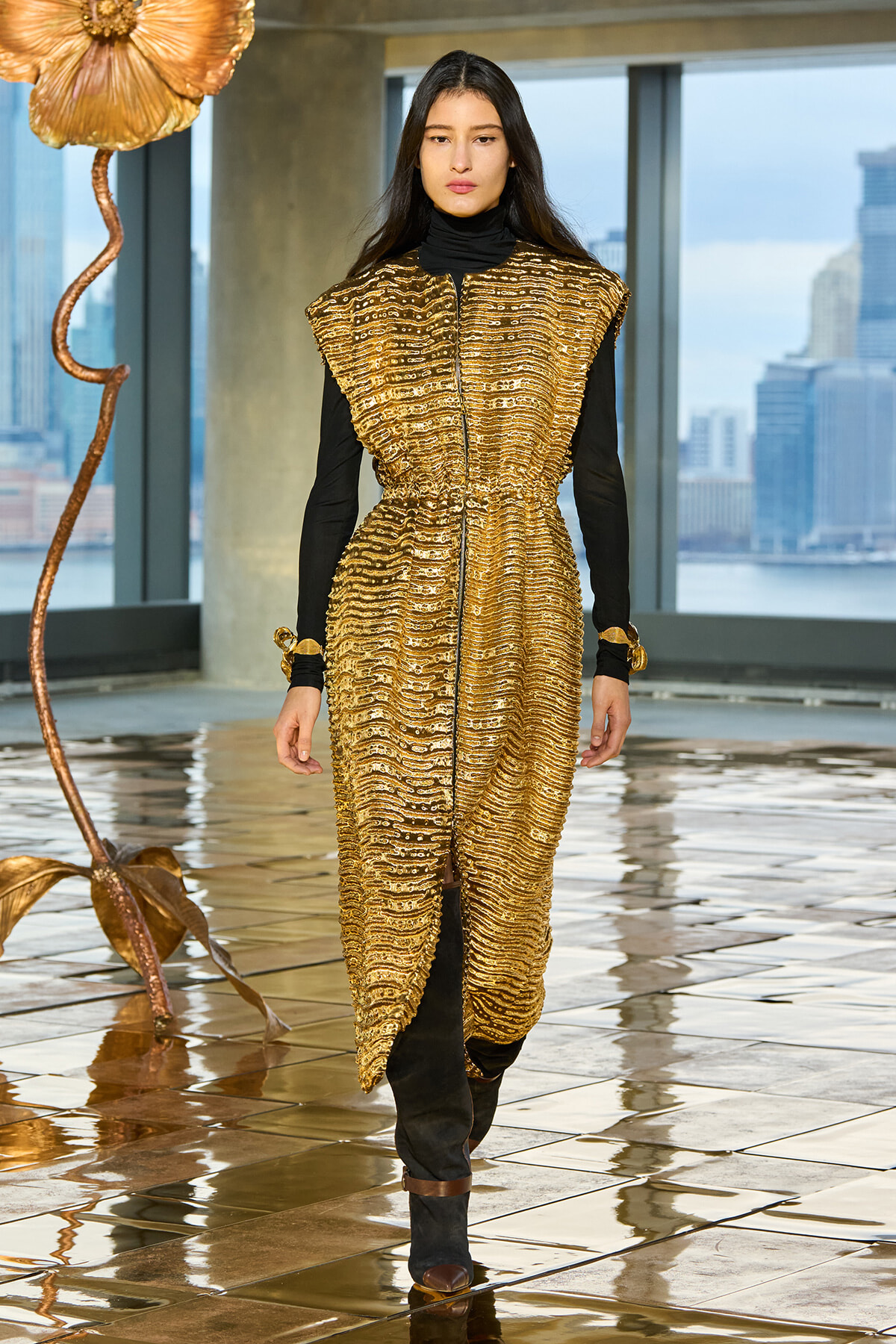 Model wearing a gold textured sleeveless coat over a black turtleneck and pants, bronze boots, walking on a reflective tiled floor with a city skyline in the background.