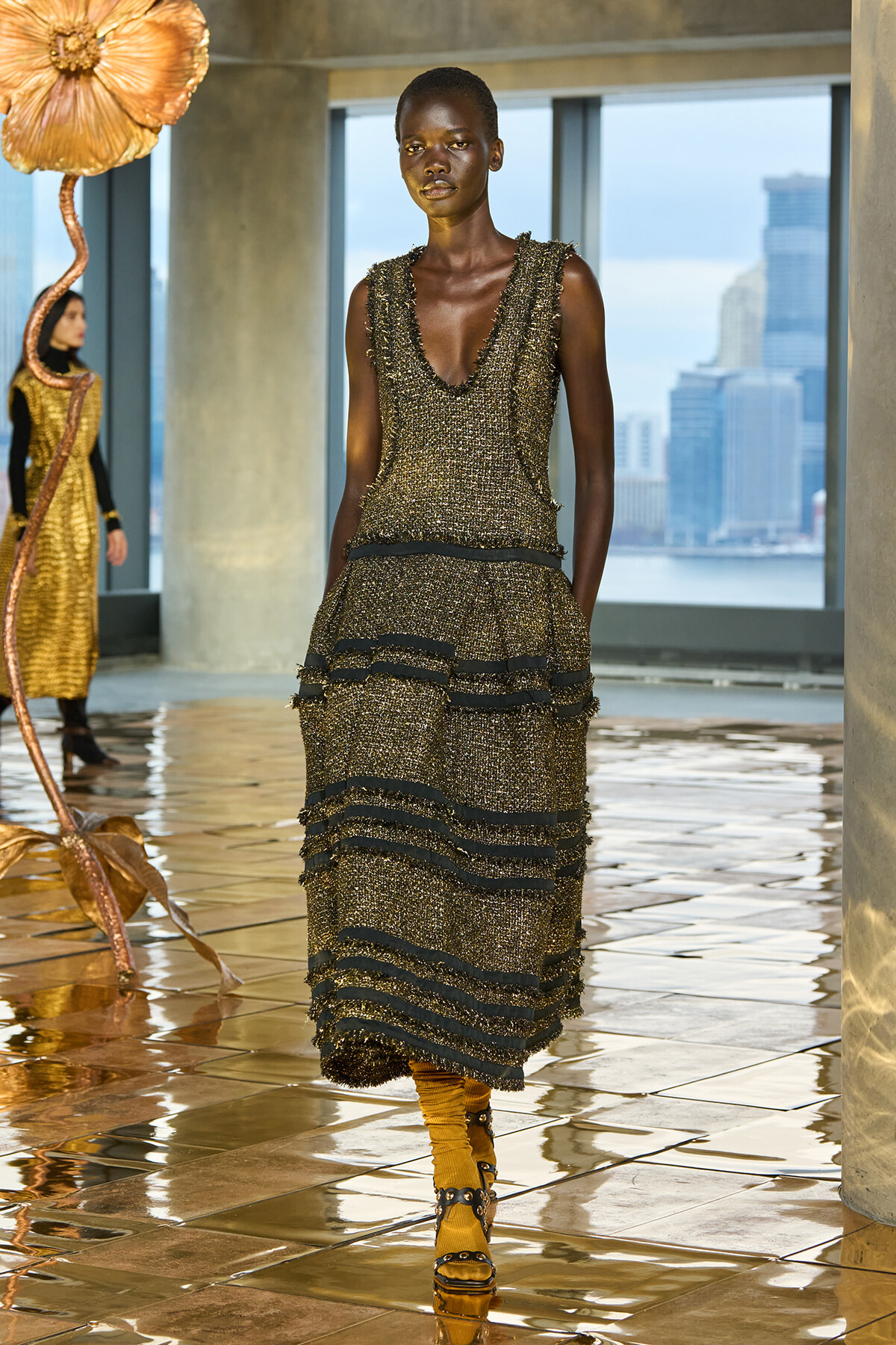 Model on a runway wearing a bronze-gold textured, sleeveless, tiered dress with black trim; city skyline visible through large windows behind.