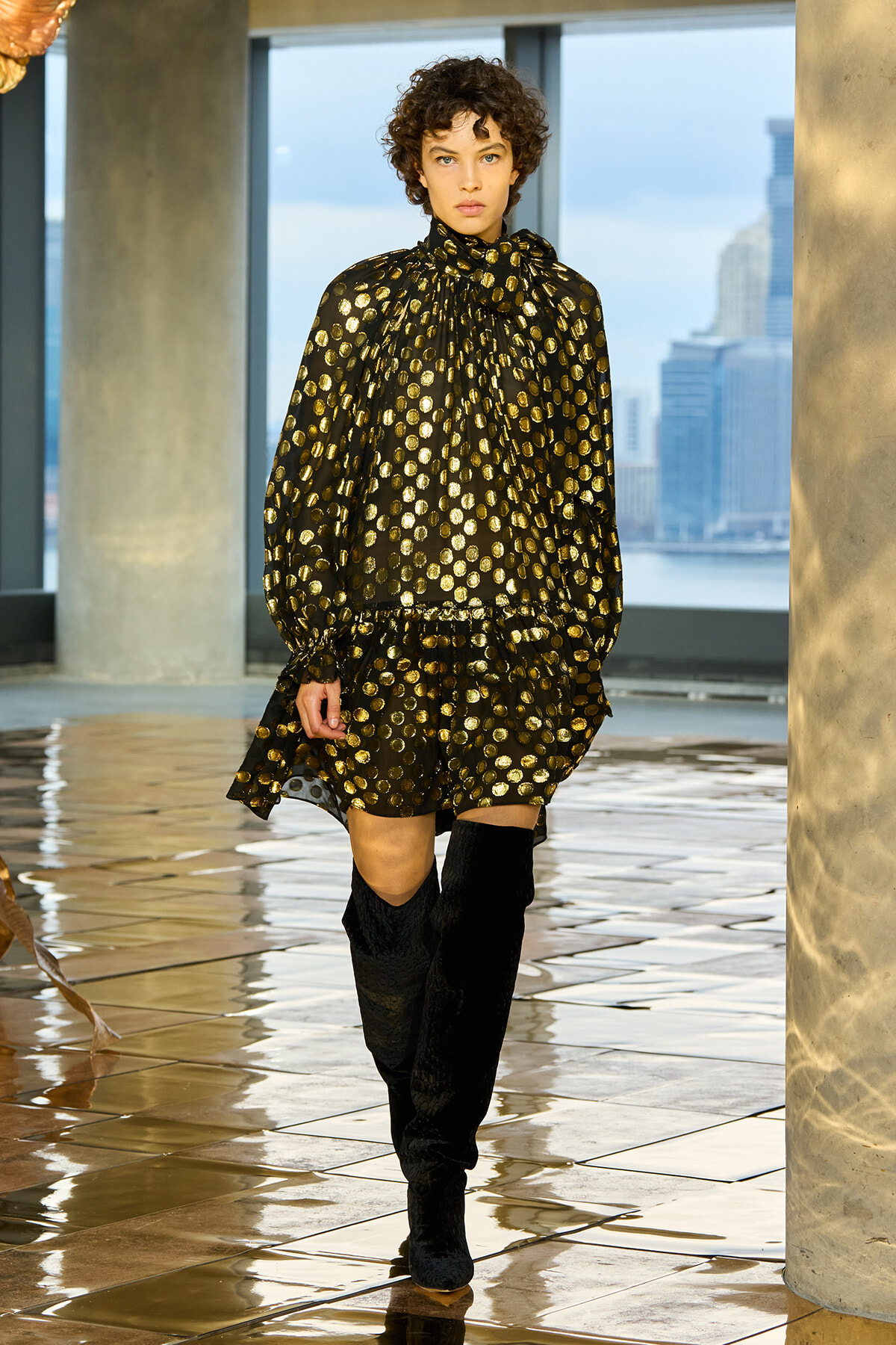 Model wearing a black dress with gold polka dots and matching knee-high black boots, walking on a glossy tile floor with a city skyline in the background.