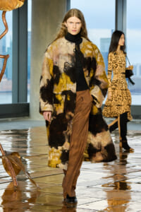 Model walks the runway in a long multicolored abstract fur coat over a black turtleneck and brown trousers; city skyline visible through windows behind.