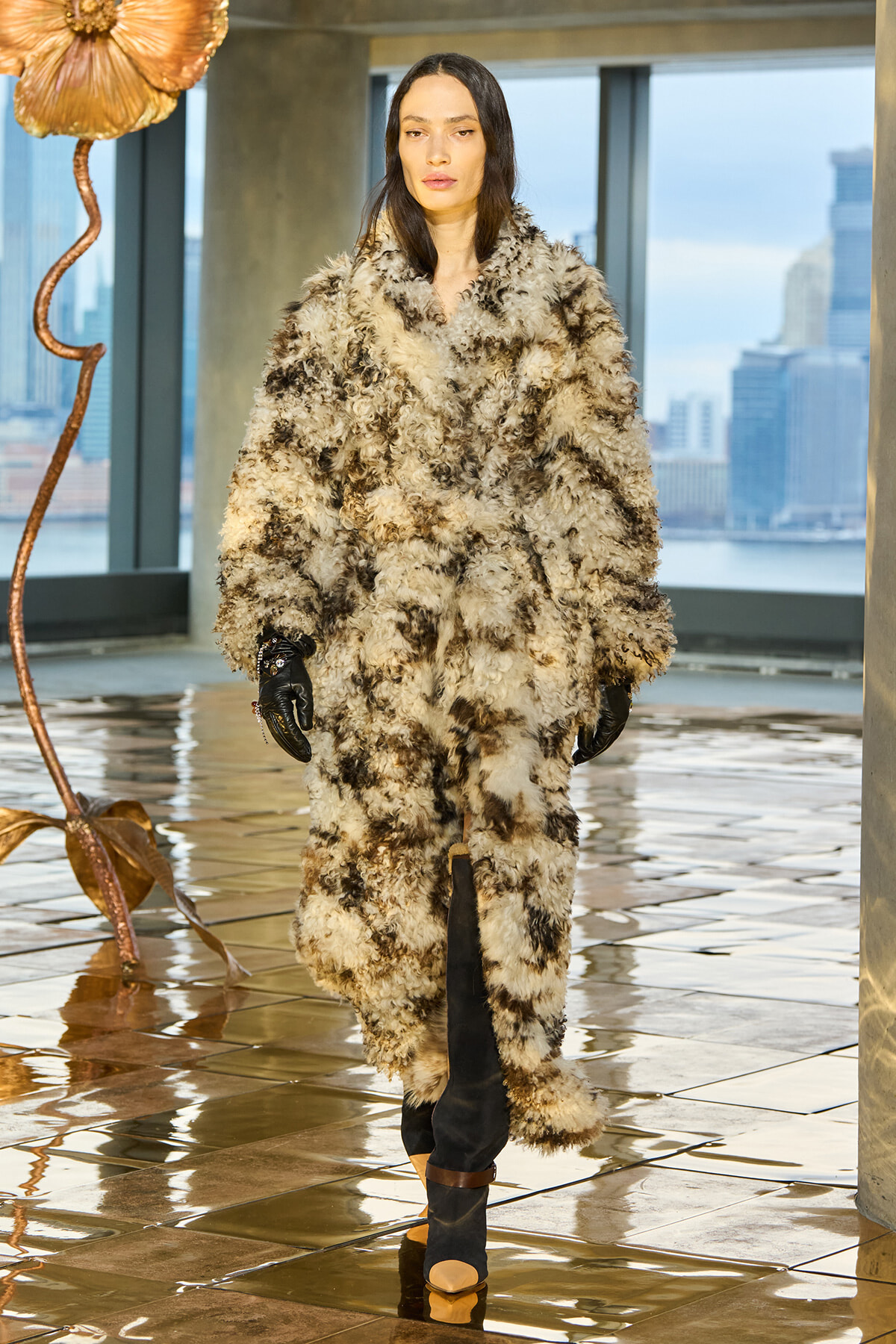 Model in a voluminous beige-brown shaggy fur coat, black gloves, and heeled boots, in a modern showroom with a city skyline.