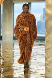 Model walking on a runway in a voluminous brown shaggy fur coat with a gold belt and green boots.