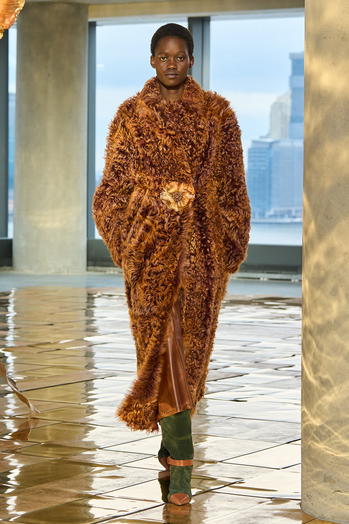 Model walking on a runway in a voluminous brown shaggy fur coat with a gold belt and green boots.
