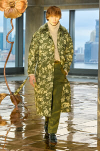 Male model on a runway wearing an olive-green floral long coat with fur collar, beige turtleneck, and olive pants; city skyline in the background.