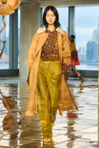 Model walking a fashion runway in a camel coat with a fur collar, sequined brown top, and olive-green leather pants.