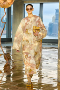Model in a pastel patchwork fur coat, oversized sunglasses, and a tan clutch walking a runway with a city skyline behind.