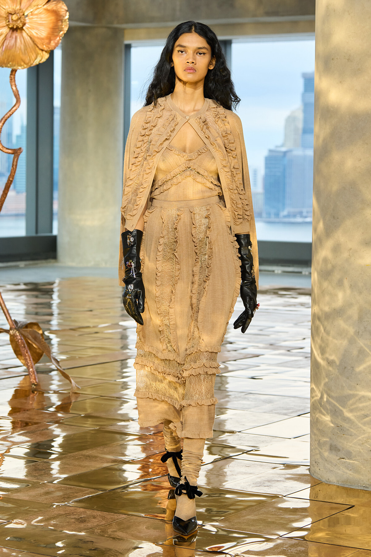 Model walking a runway in a beige, ruffled layered dress with a matching cape and black gloves, city skyline in the background