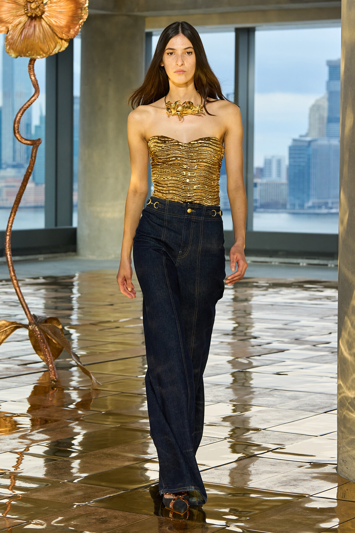 Model walks a fashion show wearing a gold strapless top with ruffles and dark wide-leg jeans, urban skyline backdrop with a copper flower sculpture nearby.