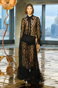 Model wearing a sheer black polka-dot gown with a bow-front collar on a reflective runway.",