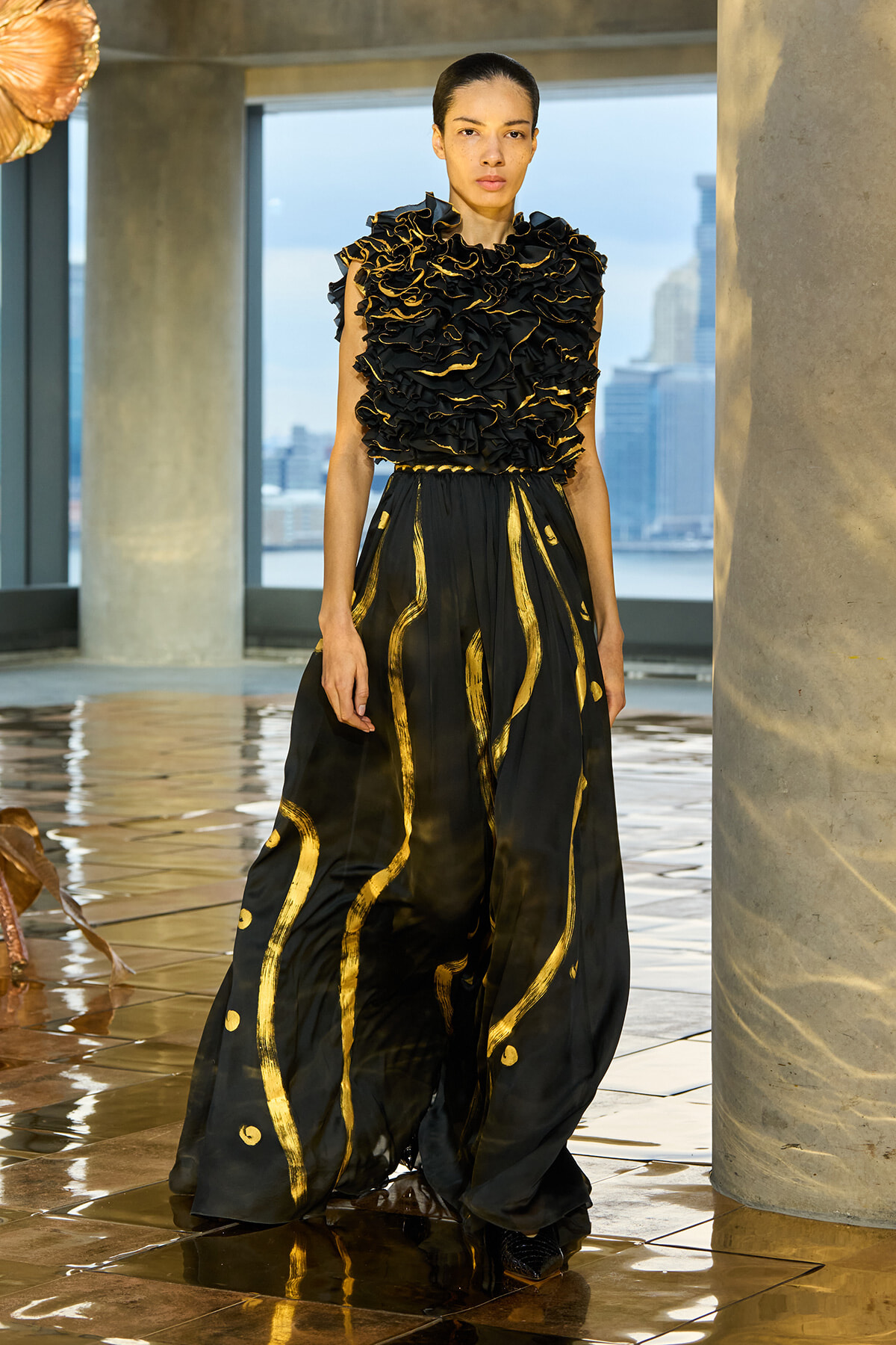 Model wearing a black ball gown with a heavily ruffled bodice and gold piping, standing in a modern, sunlit loft with city skyline outside.