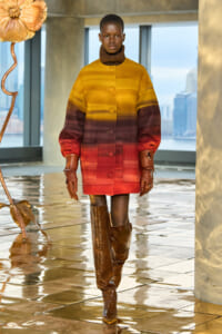 Model wearing a yellow-to-red gradient oversized coat, brown turtleneck, and knee-high crocodile-embossed boots indoors with city skyline in the background.