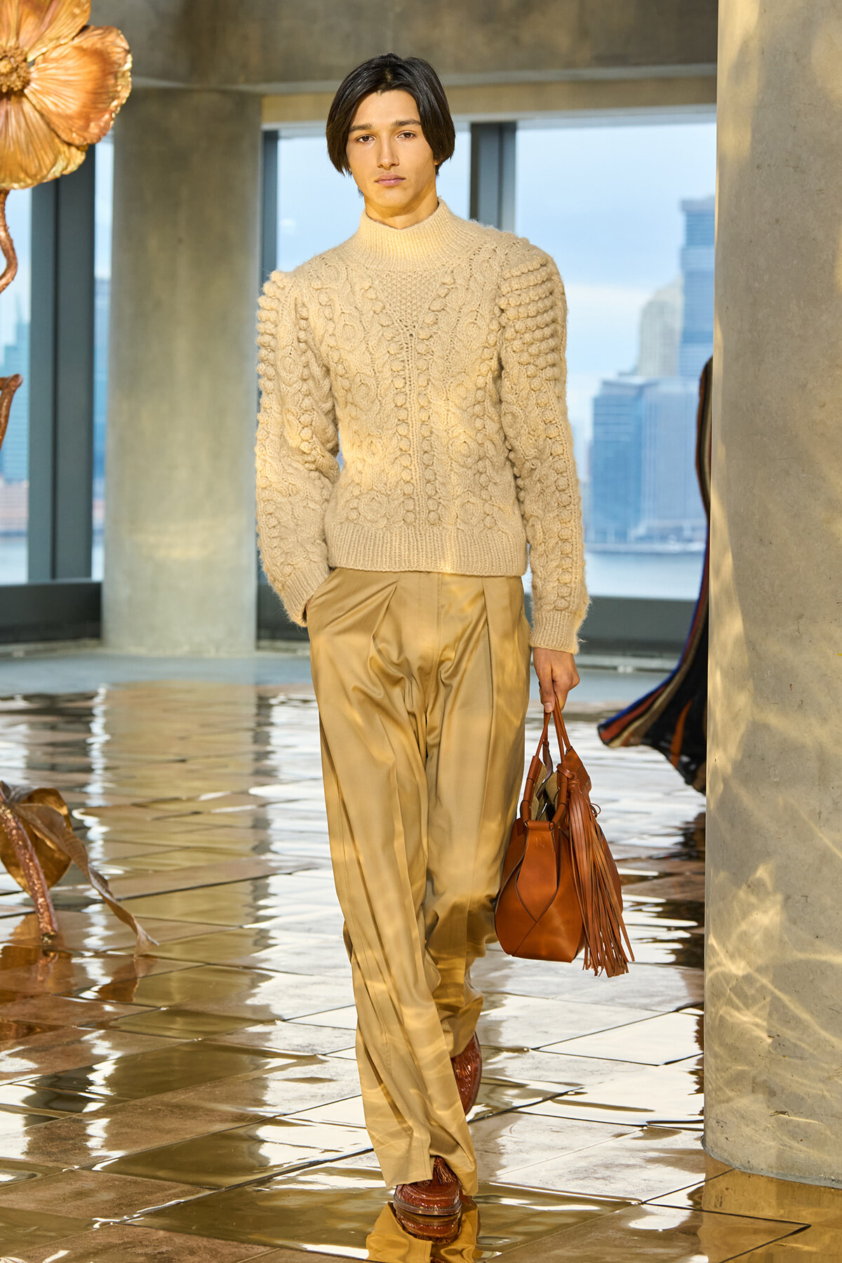 Model walking a runway in a cream cable-knit sweater and gold satin trousers, carrying a brown fringe handbag.