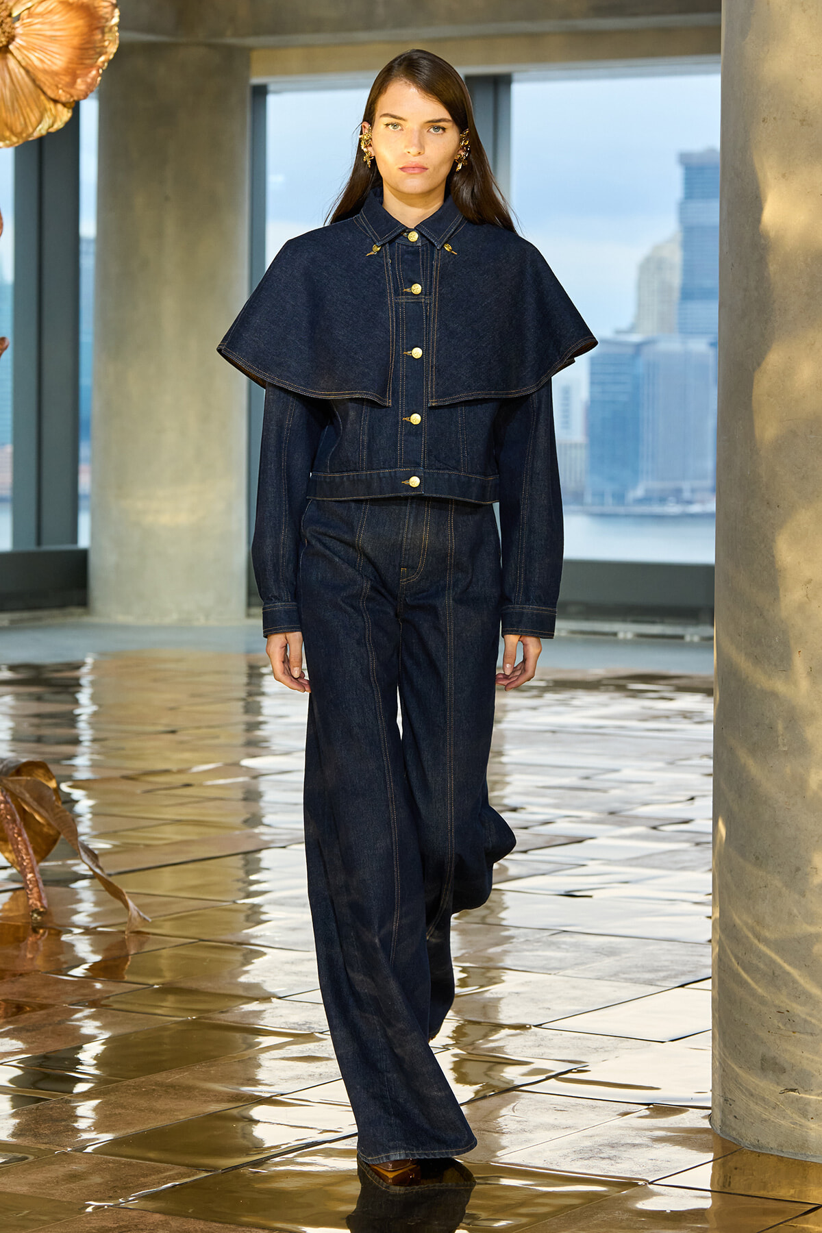 Female model in a dark denim cape jacket with gold buttons over matching wide-leg jeans, in an urban loft setting with a city skyline.