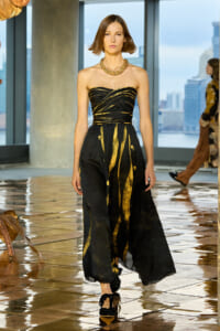 Model walks a runway in a strapless black gown with gold accents and a chunky gold necklace, against a city skyline backdrop.