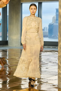 Model walking runway in a cream sheer gown with ruffled trim and long sleeves, city skyline in the background.